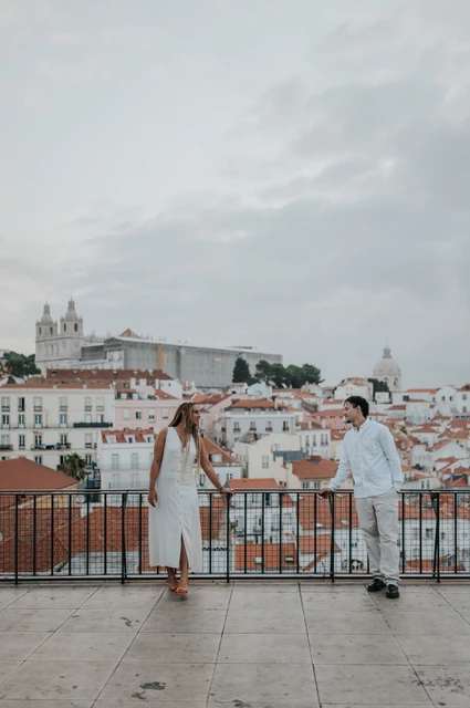 Hand in hand above Lisbon’s rooftops | Lisbon couple photoshoot The couple holding hands beside the viewpoint railing with Lisbon’s rooftops behind them during their couple photoshoot in Lisbon.