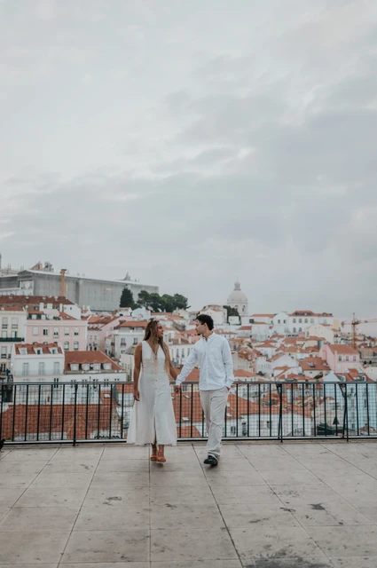 Hand in hand above Lisbon’s rooftops | Lisbon couple photoshoot The couple holding hands beside the viewpoint railing with Lisbon’s rooftops behind them during their couple photoshoot in Lisbon.