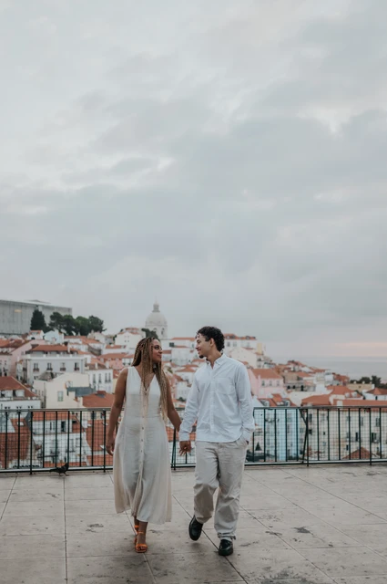 Hand in hand above Lisbon’s rooftops | Lisbon couple photoshoot The couple holding hands beside the viewpoint railing with Lisbon’s rooftops behind them during their couple photoshoot in Lisbon.