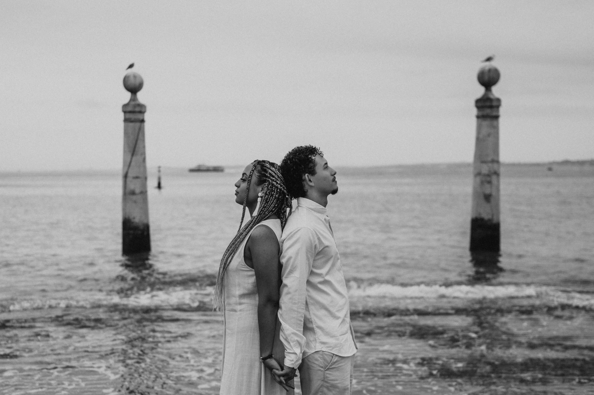 Black-and-white photograph: the couple standing back to back on the waterfront steps beside the Tagus River during their couple photoshoot in Lisbon.