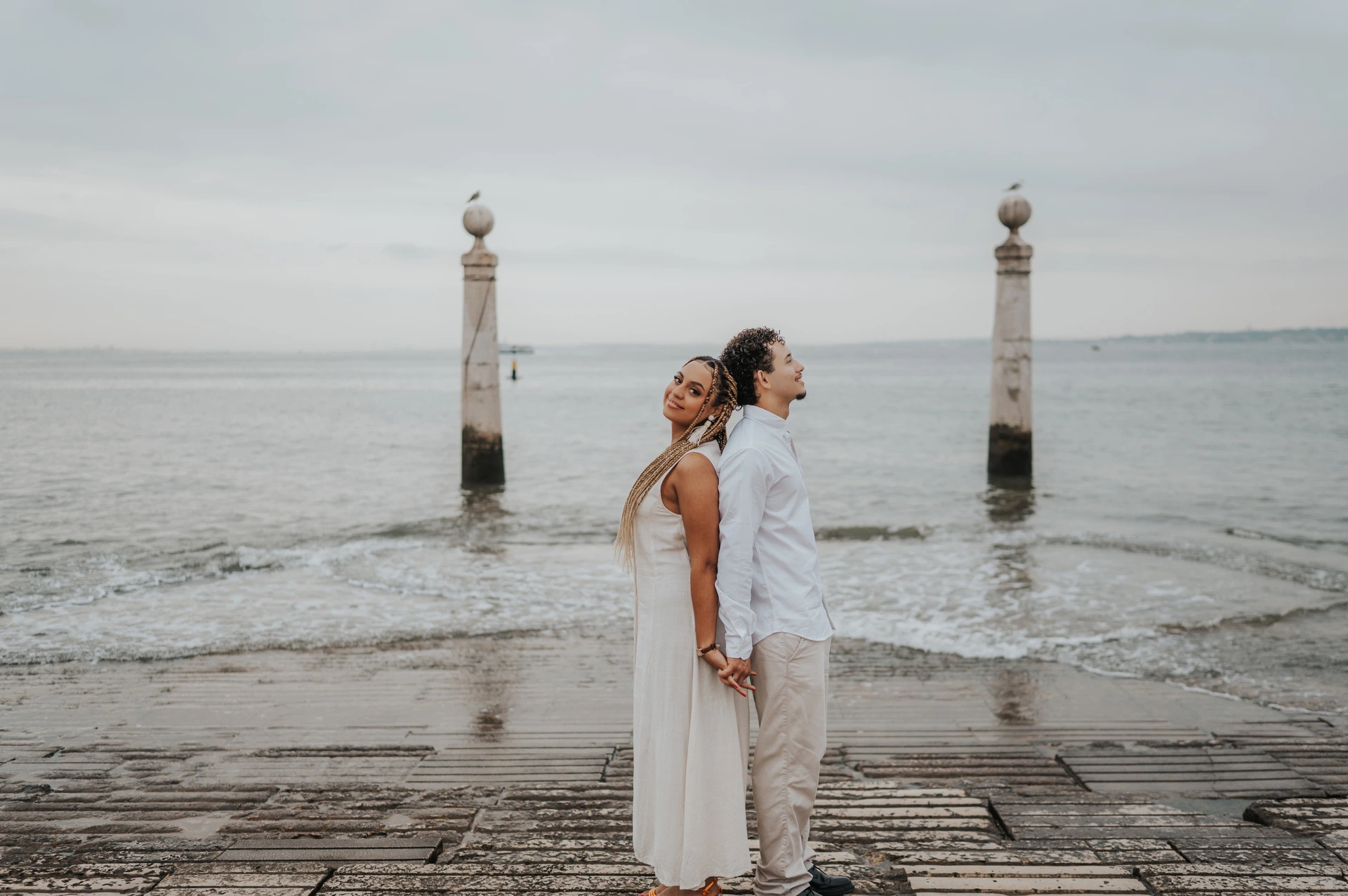 The couple standing back to back on the waterfront steps beside the Tagus River during their couple photoshoot in Lisbon.