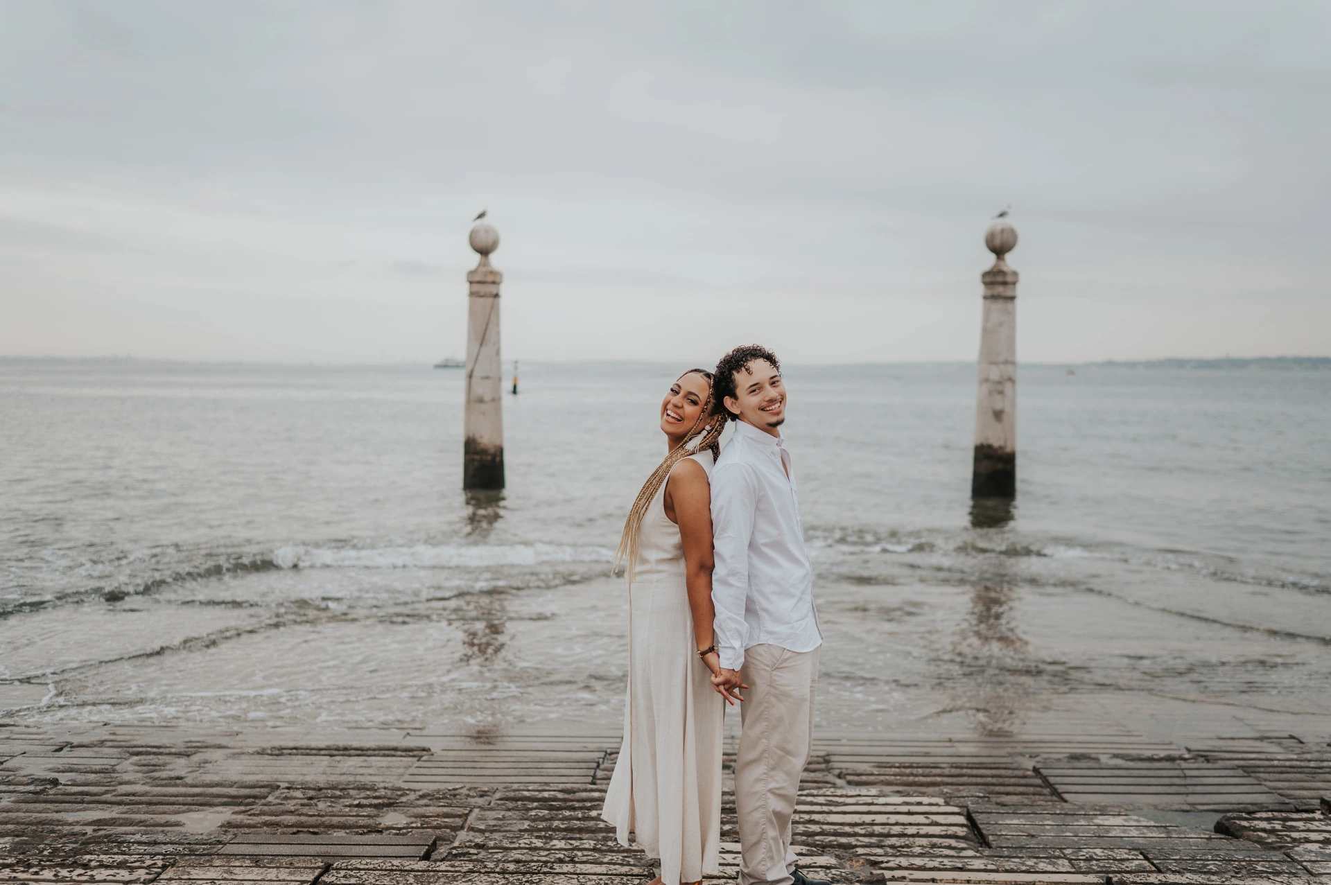 The couple standing back to back on the waterfront steps beside the Tagus River during their couple photoshoot in Lisbon.