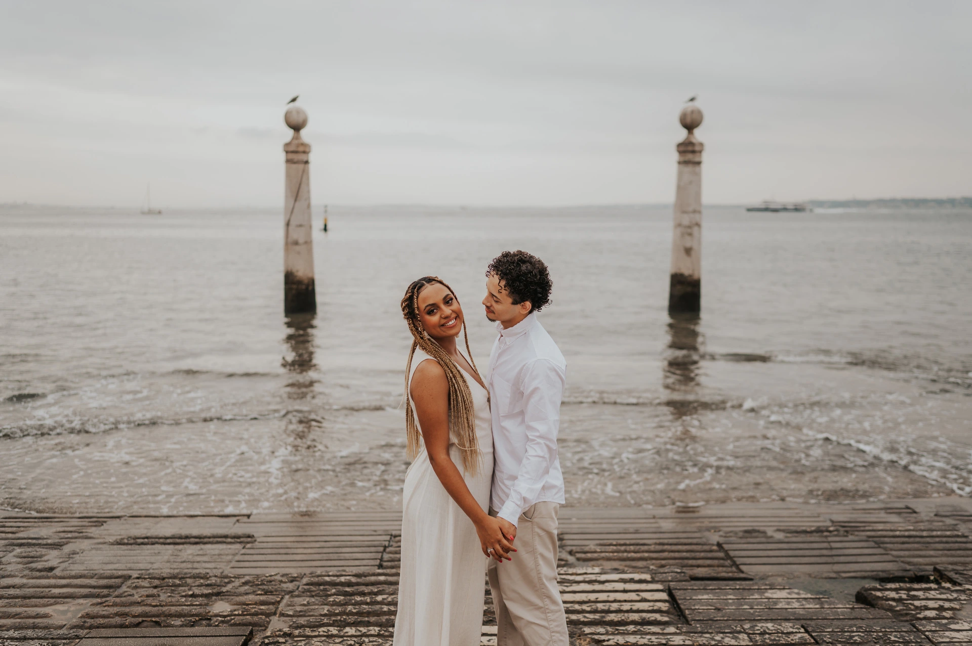 The couple standing close together on the waterfront steps with the river behind them during their couple photoshoot in Lisbon.