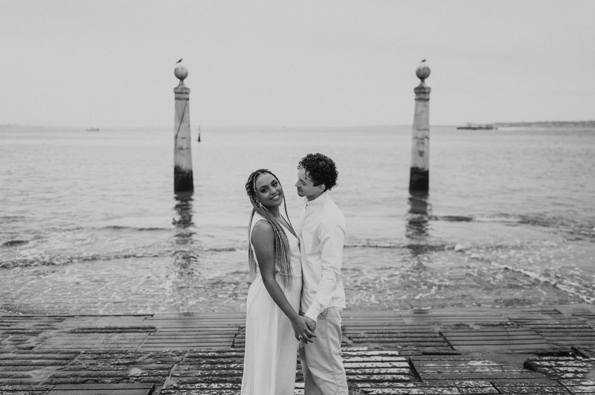 Black-and-white photograph: the couple standing close together on the waterfront steps with the river behind them during their couple photoshoot in Lisbon.
