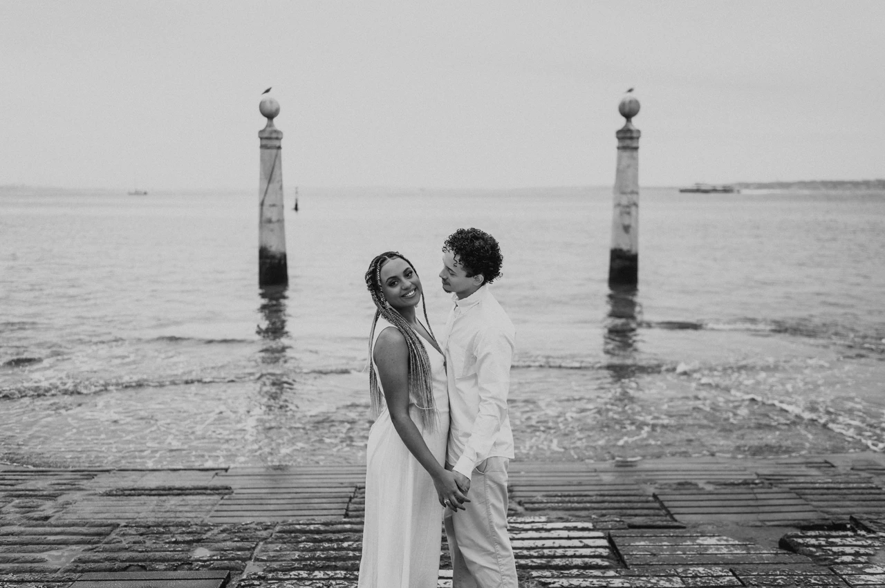 Close together by the water in black and white | Lisbon couple photoshoot Black-and-white photograph: the couple standing close together on the waterfront steps with the river behind them during their couple photoshoot in Lisbon.