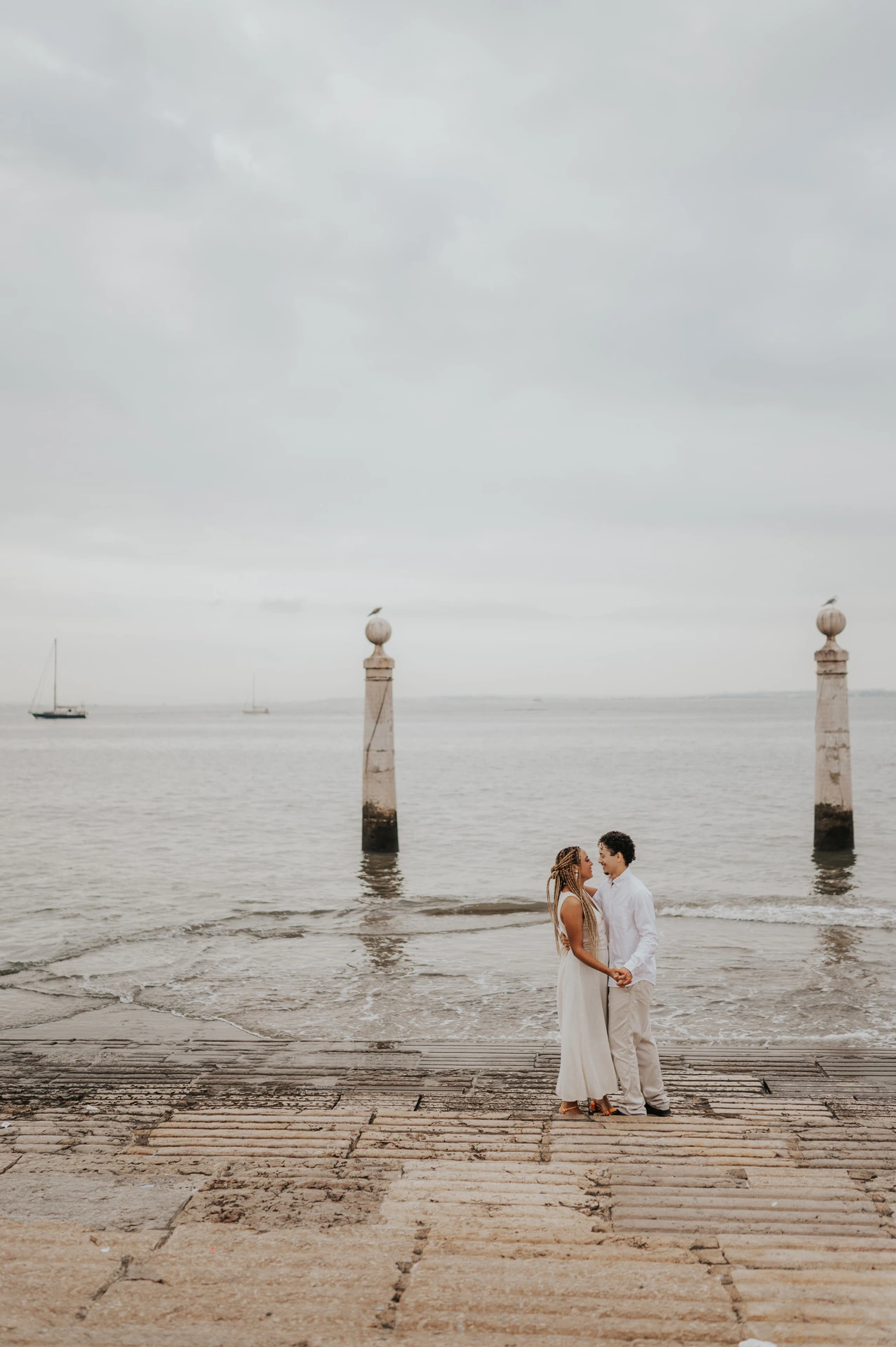 The couple kissing on the waterfront steps with the river and two stone columns behind them during their couple photoshoot in Lisbon.