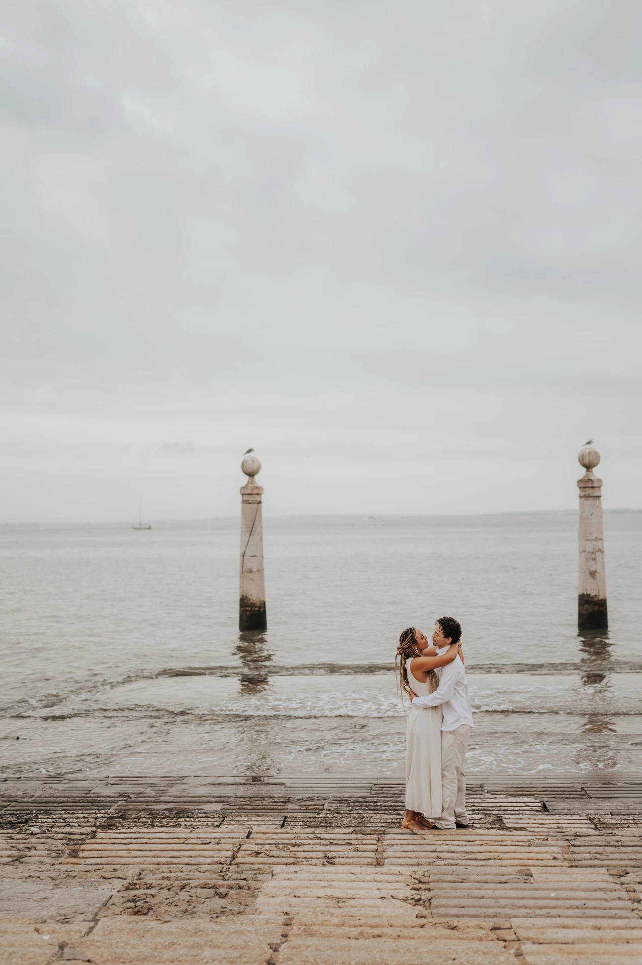 The couple kissing on the waterfront steps with the river and two stone columns behind them during their couple photoshoot in Lisbon.