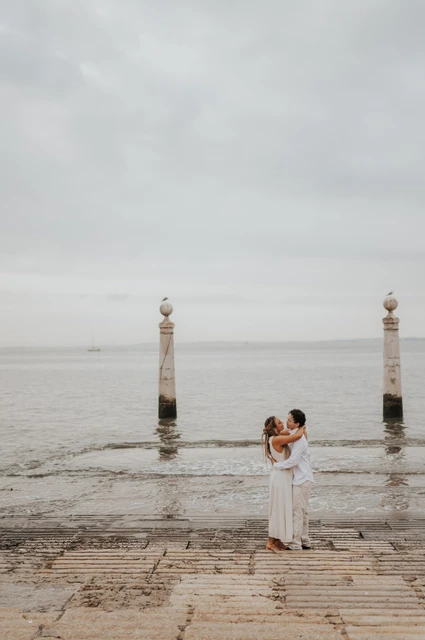 A kiss beside the Tagus River | Lisbon couple photoshoot The couple kissing on the waterfront steps with the river and two stone columns behind them during their couple photoshoot in Lisbon.