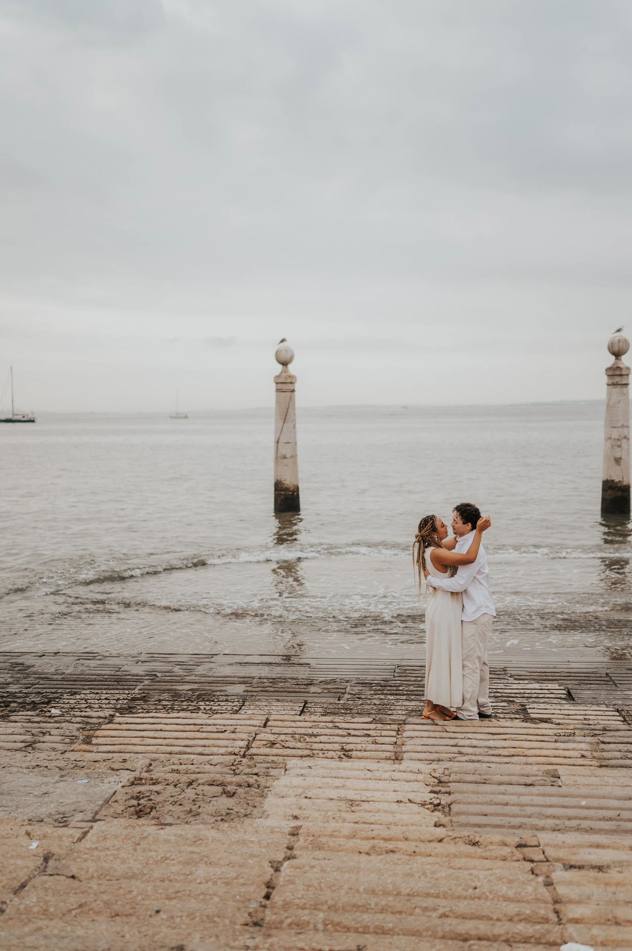The couple kissing on the waterfront steps with the river and two stone columns behind them during their couple photoshoot in Lisbon.