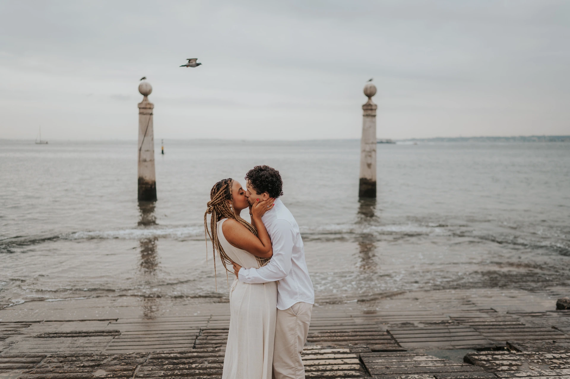 The couple kissing on the waterfront steps with the river and two stone columns behind them during their couple photoshoot in Lisbon.