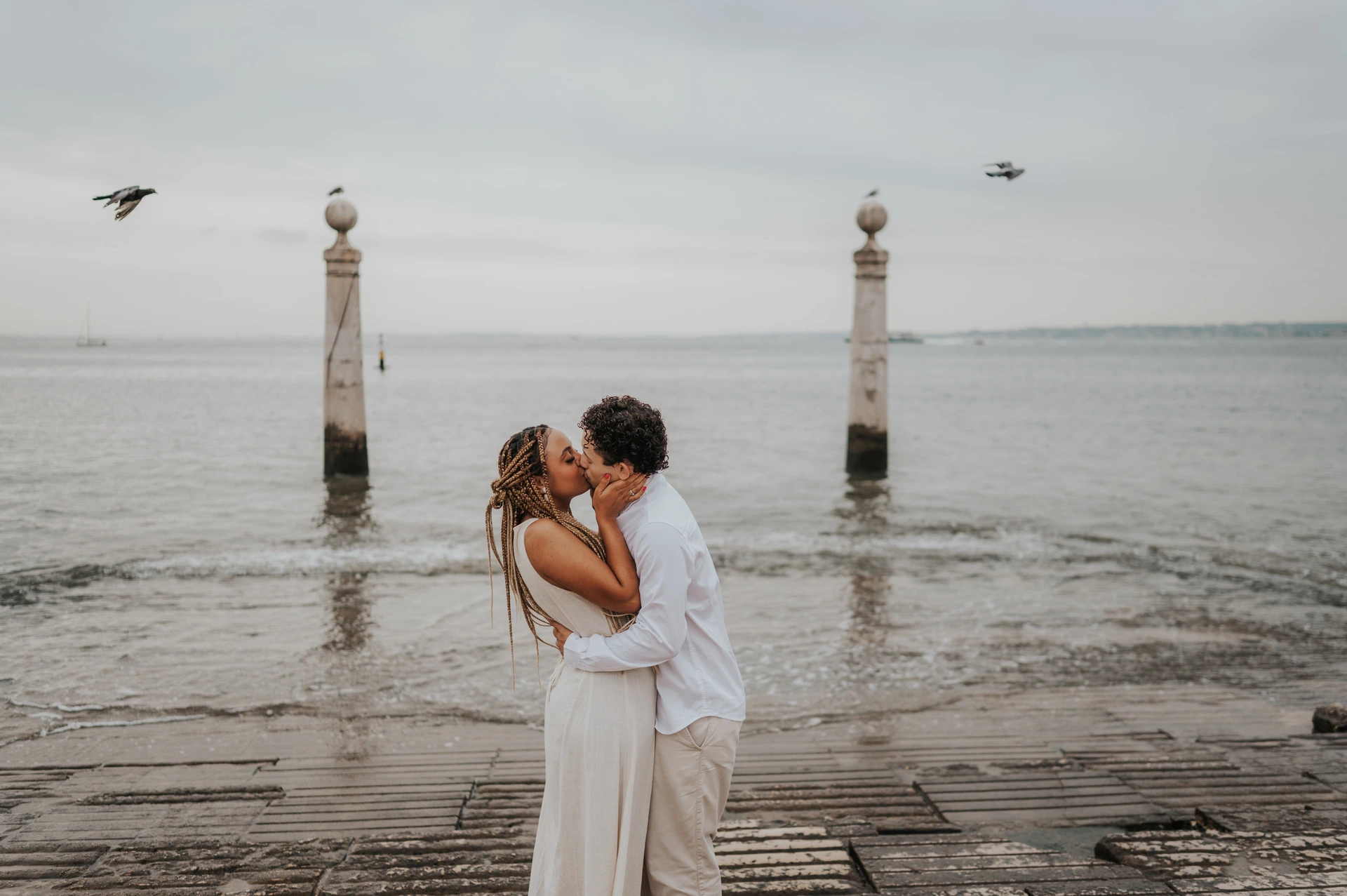 The couple kissing on the waterfront steps with the river and two stone columns behind them during their couple photoshoot in Lisbon.