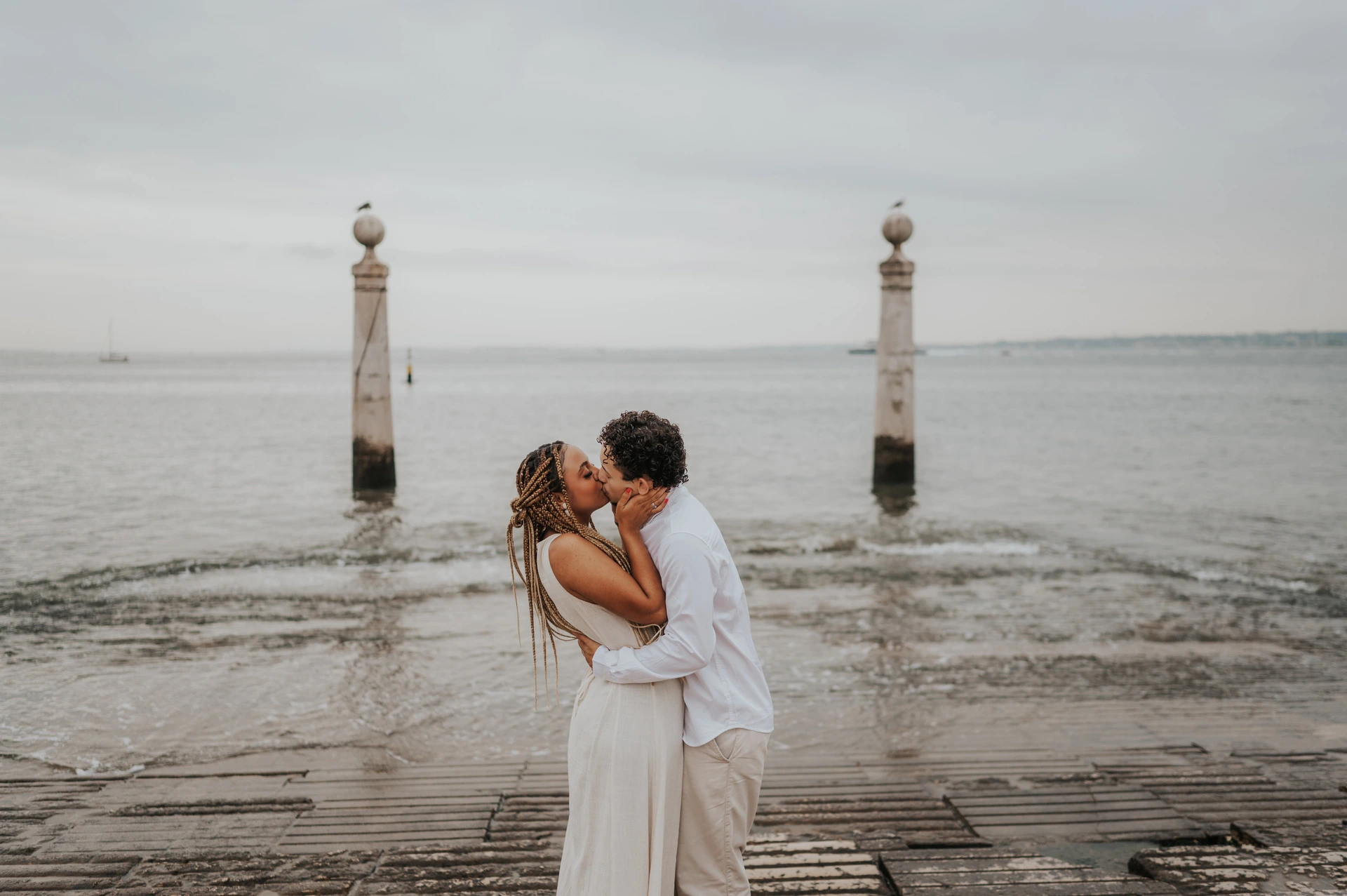 The couple kissing on the waterfront steps with the river and two stone columns behind them during their couple photoshoot in Lisbon.