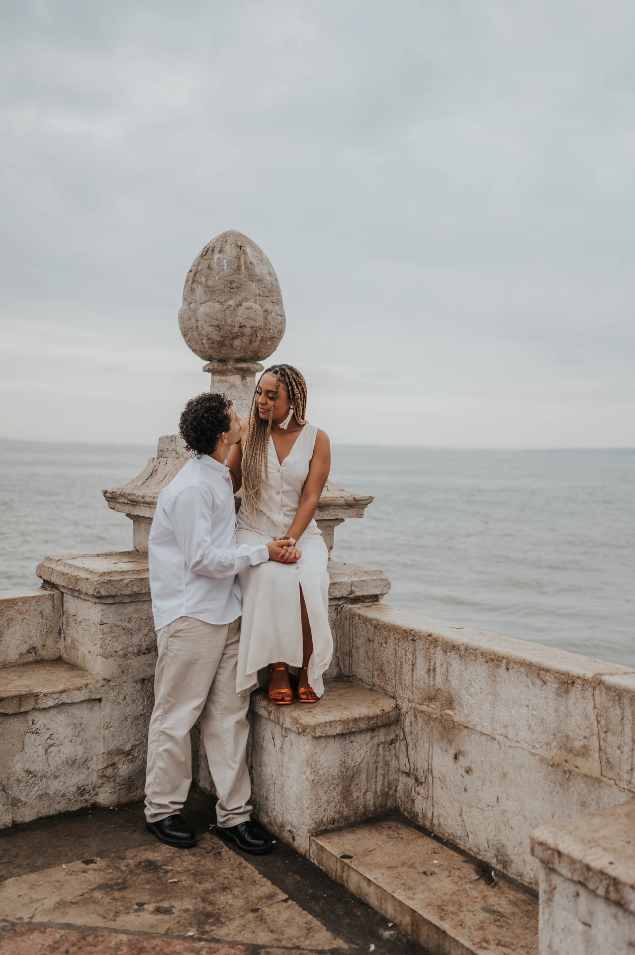 The woman seated on the low stone waterfront wall with her partner close beside her during their couple photoshoot in Lisbon.