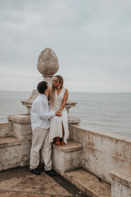 A seated portrait at the stone waterfront wall | Lisbon couple photoshoot The woman seated on the low stone waterfront wall with her partner close beside her during their couple photoshoot in Lisbon.