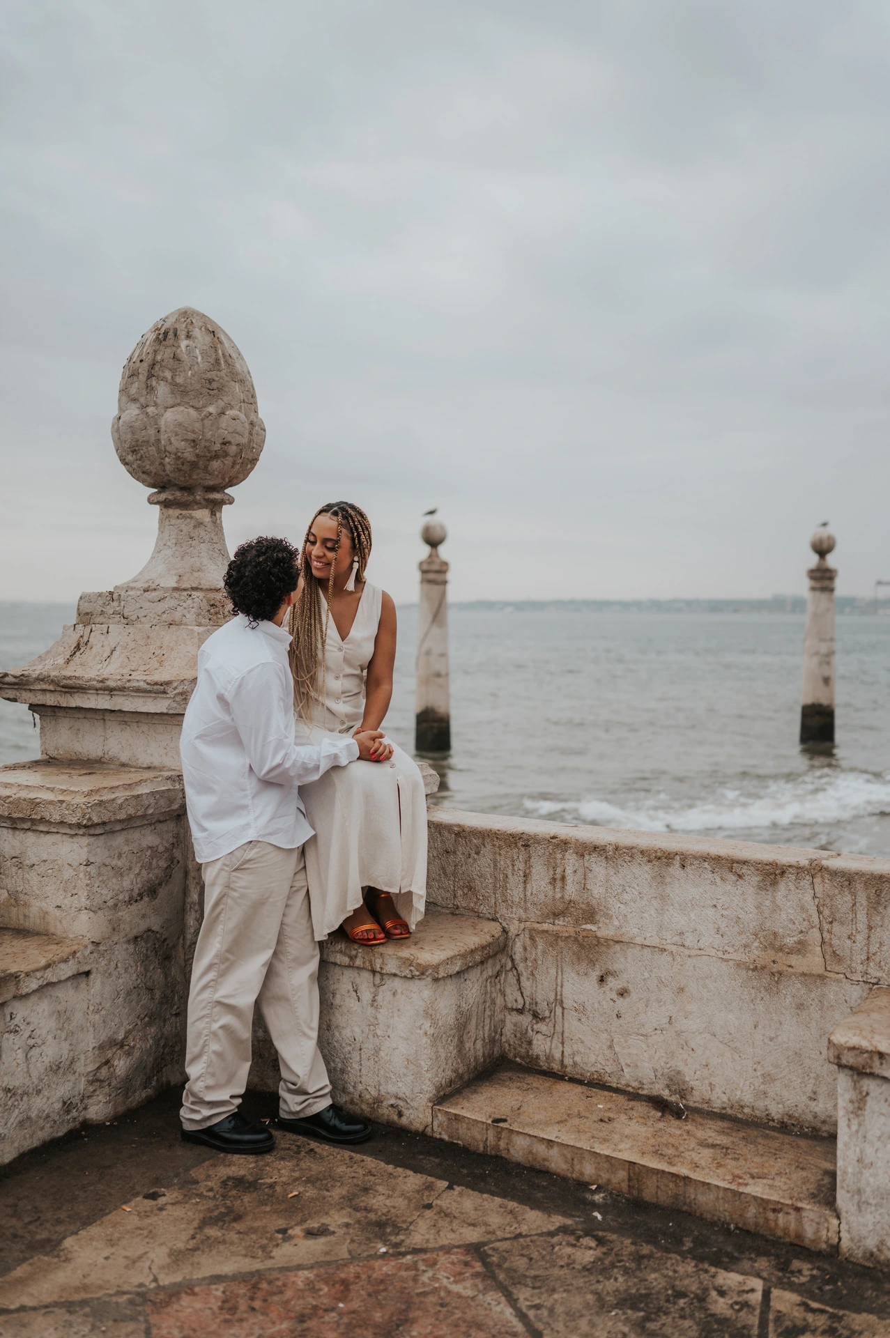The woman seated on the low stone waterfront wall with her partner close beside her during their couple photoshoot in Lisbon.