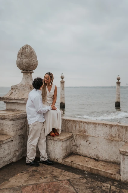 The woman seated on the low stone waterfront wall with her partner close beside her during their couple photoshoot in Lisbon.