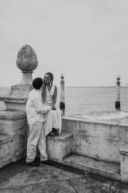 A seated portrait at the stone waterfront wall in black and white | Lisbon couple photoshoot Black-and-white photograph: the woman seated on the low stone waterfront wall with her partner close beside her during their couple photoshoot in Lisbon.