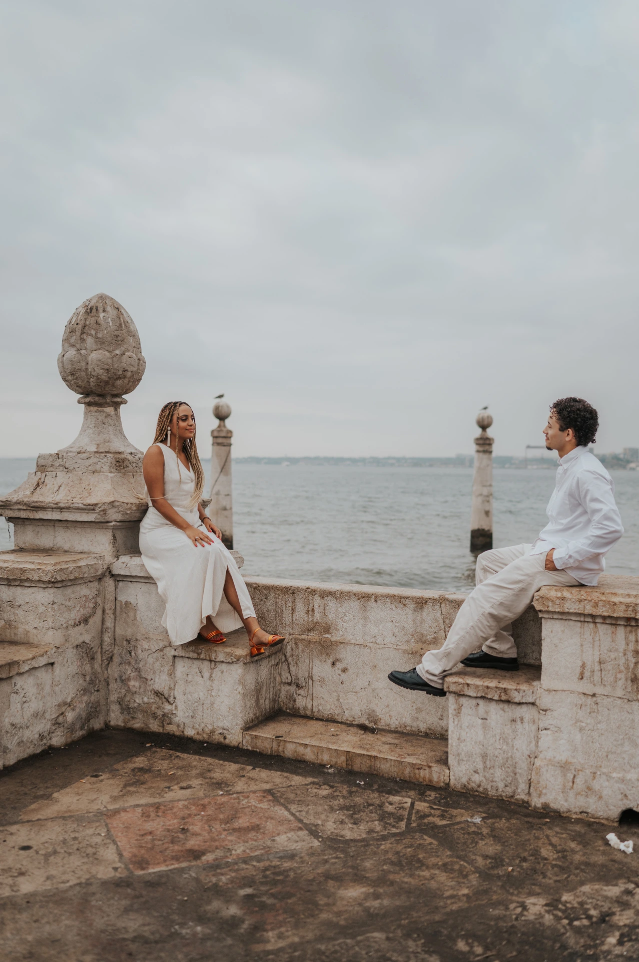 The woman seated on the low stone waterfront wall with her partner close beside her during their couple photoshoot in Lisbon.