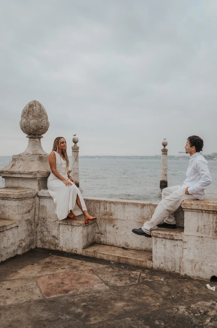 A seated portrait at the stone waterfront wall | Lisbon couple photoshoot The woman seated on the low stone waterfront wall with her partner close beside her during their couple photoshoot in Lisbon.