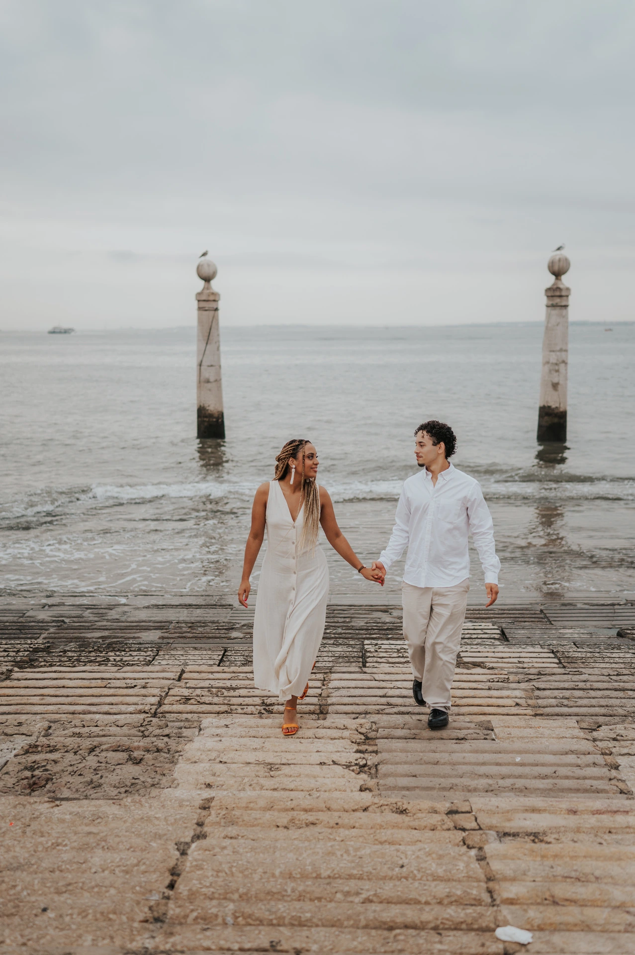 The couple smiling and holding hands as they walk up the stone waterfront steps during their couple photoshoot in Lisbon.