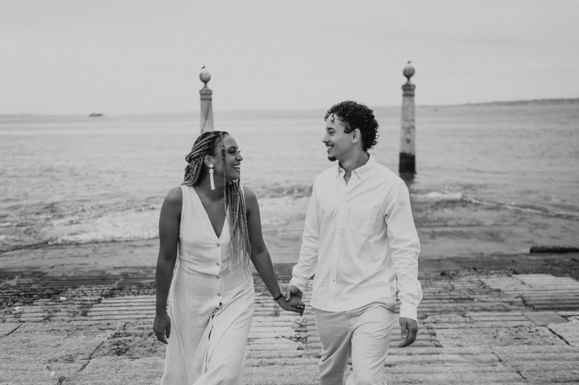 Black-and-white photograph: the couple smiling and holding hands as they walk up the stone waterfront steps during their couple photoshoot in Lisbon.