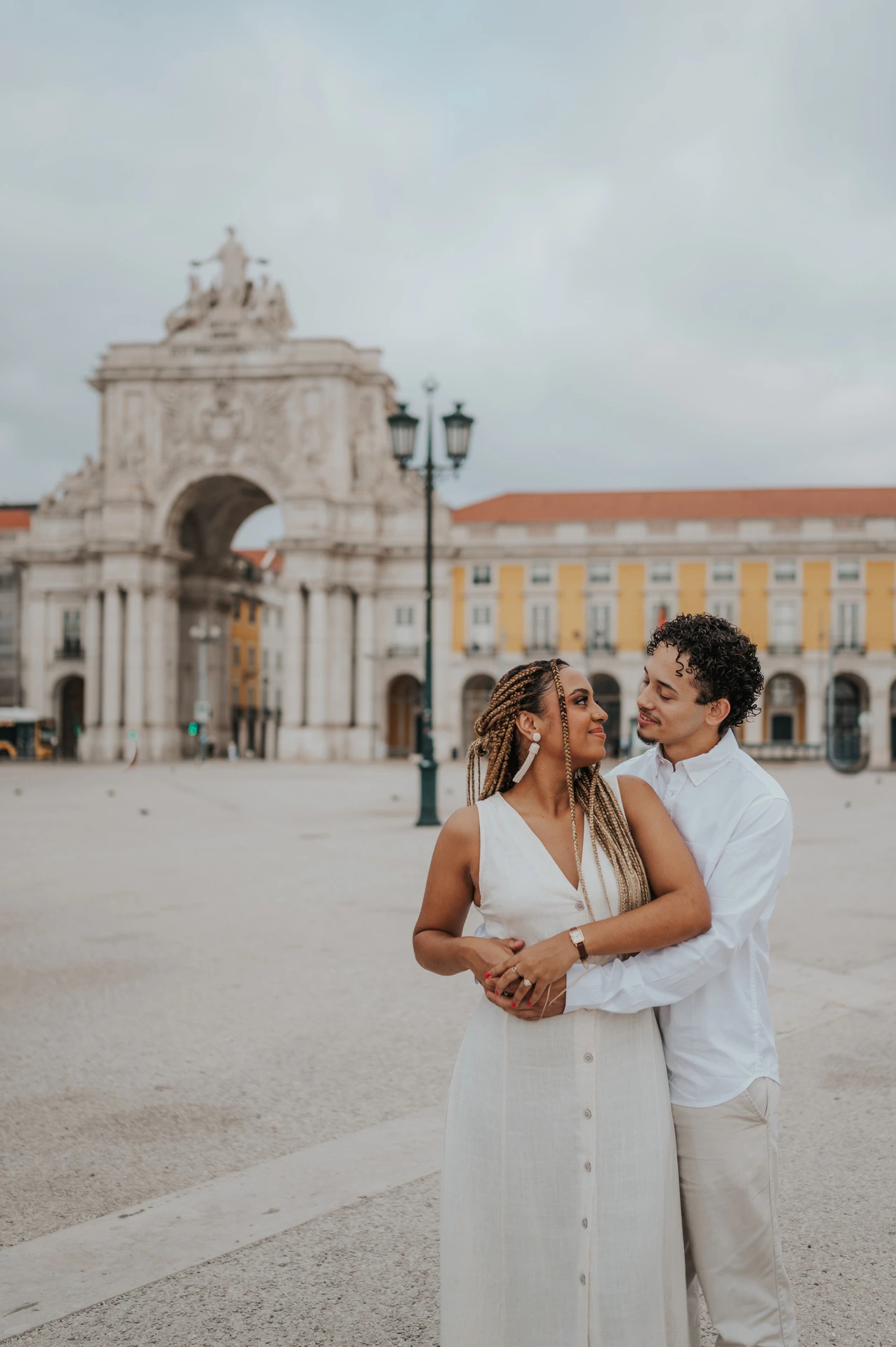 The man embracing his smiling partner from behind with Praça do Comércio in the background during their couple photoshoot in Lisbon.