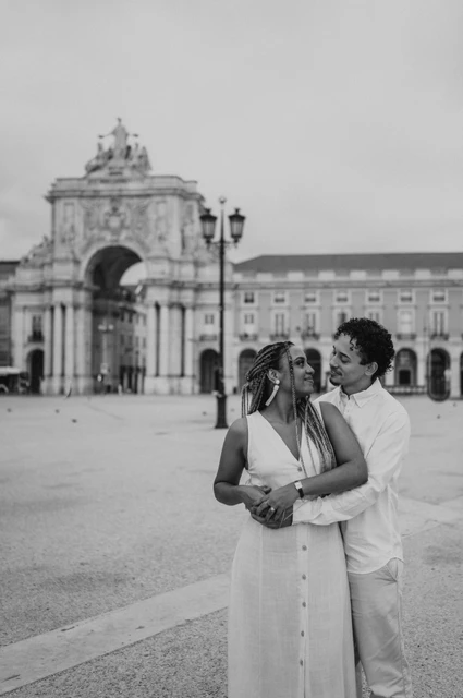 An embrace in Praça do Comércio in black and white | Lisbon couple photoshoot Black-and-white photograph: the man embracing his smiling partner from behind with Praça do Comércio in the background during their couple photoshoot in Lisbon.