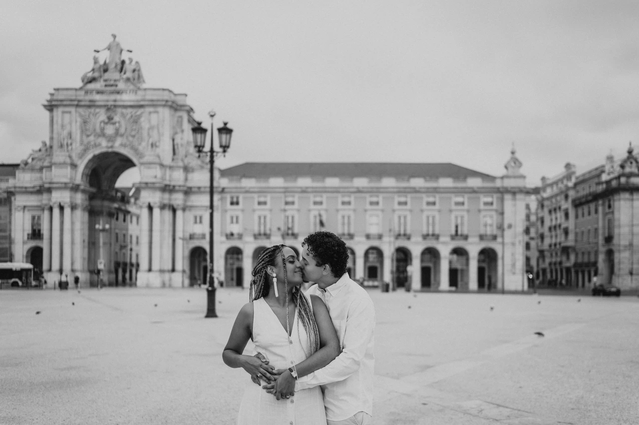 An embrace in Praça do Comércio in black and white | Lisbon couple photoshoot Black-and-white photograph: the man embracing his smiling partner from behind with Praça do Comércio in the background during their couple photoshoot in Lisbon.
