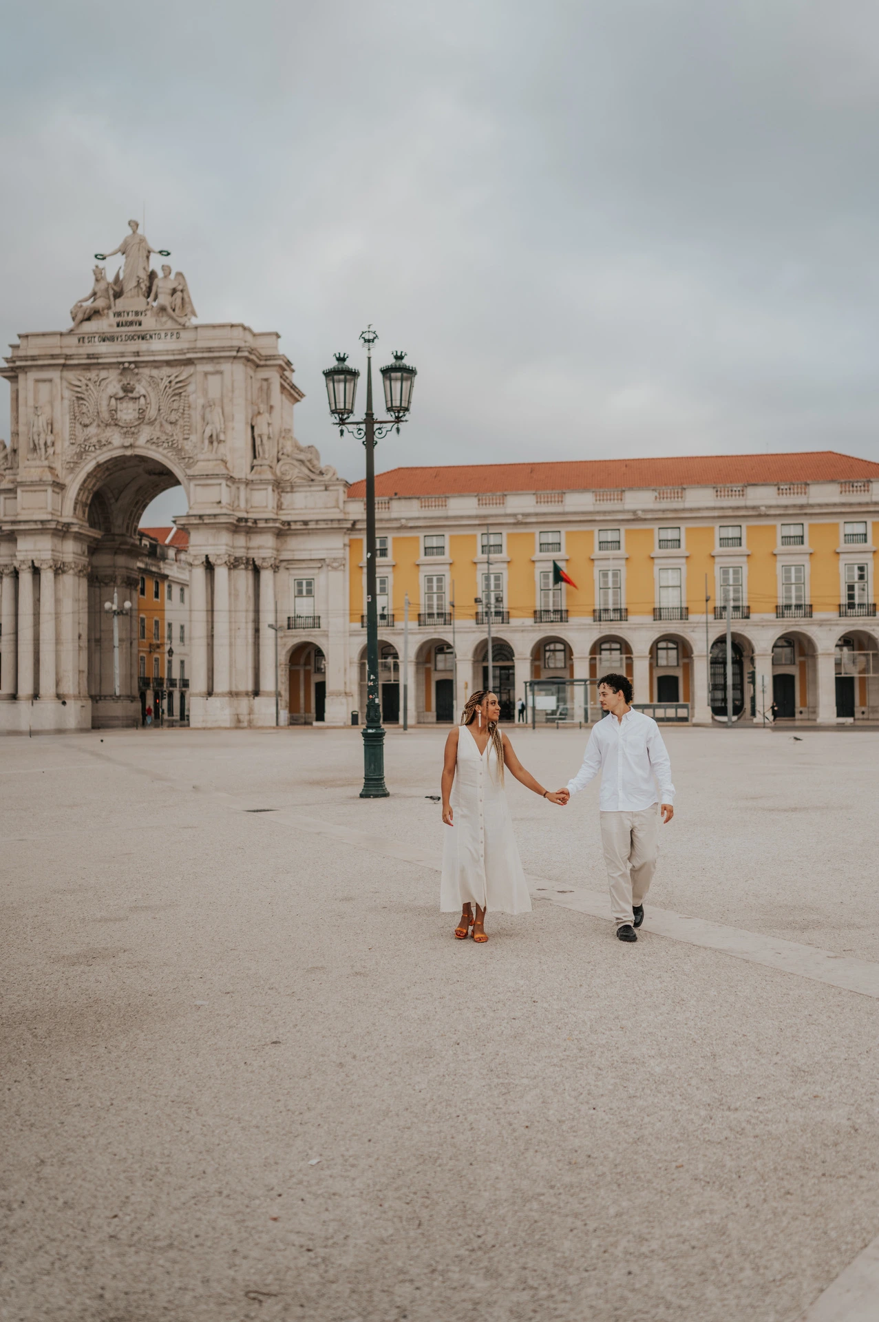 The couple walking hand in hand across Praça do Comércio with the monumental arch behind them during their couple photoshoot in Lisbon.