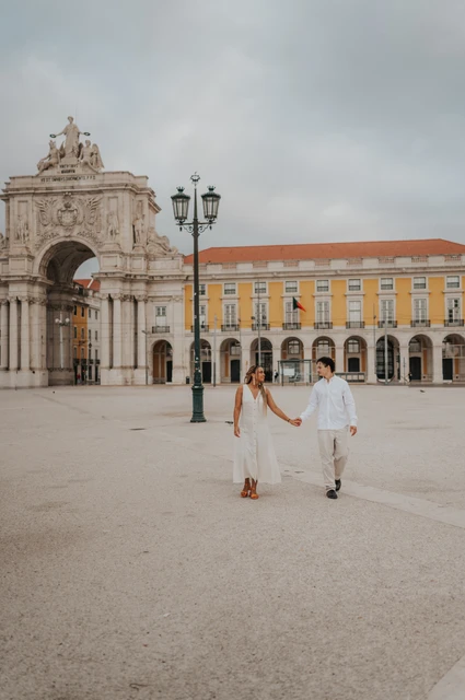 The couple walking hand in hand across Praça do Comércio with the monumental arch behind them during their couple photoshoot in Lisbon.