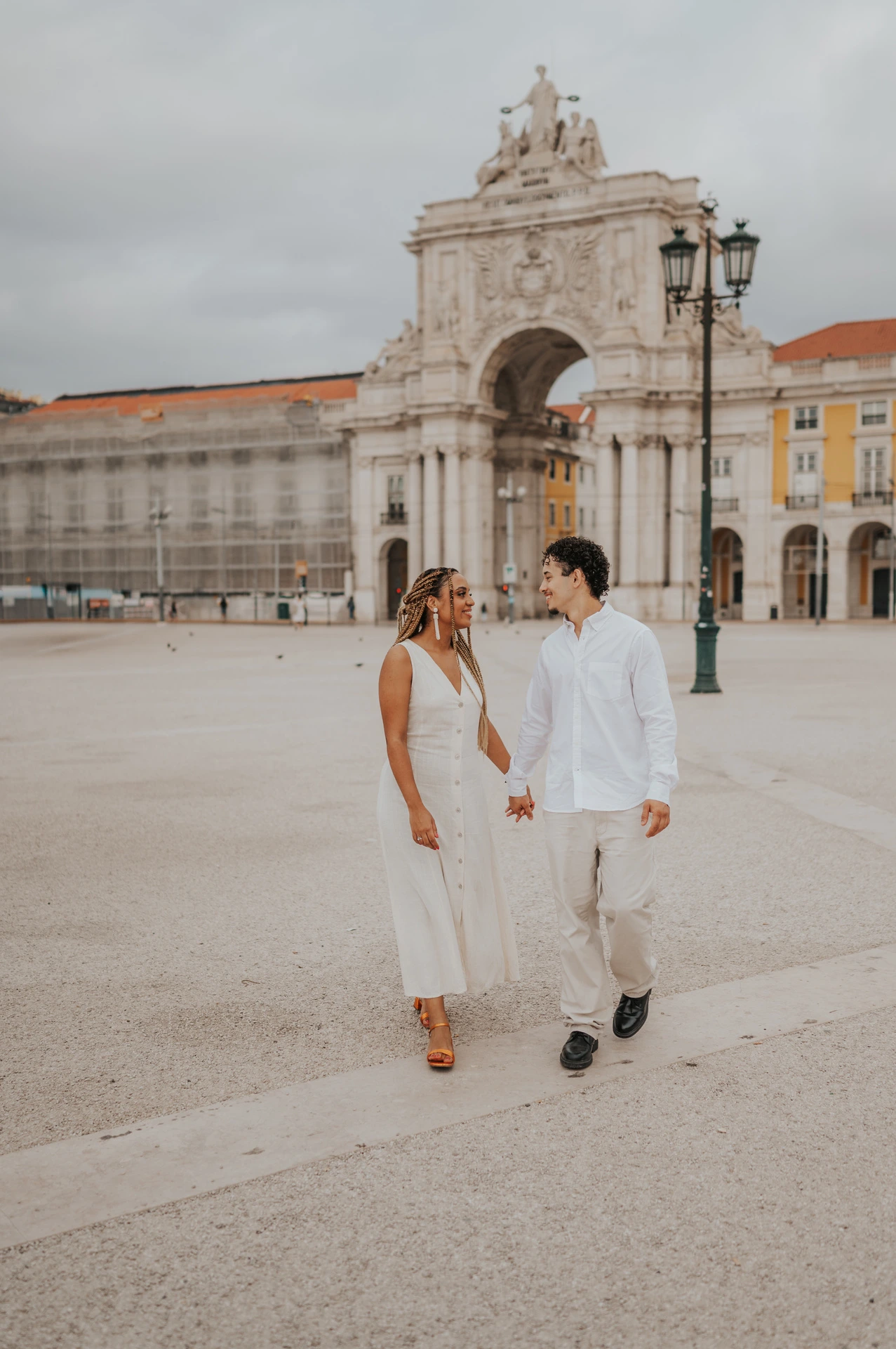 The couple walking hand in hand across Praça do Comércio with the monumental arch behind them during their couple photoshoot in Lisbon.