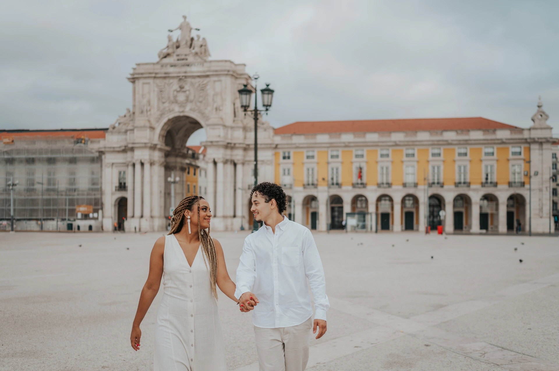 The couple walking hand in hand across Praça do Comércio with the monumental arch behind them during their couple photoshoot in Lisbon.