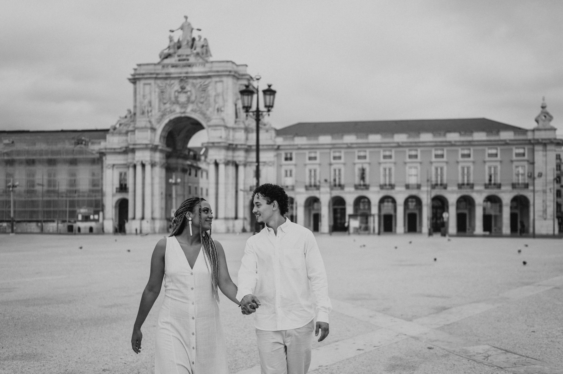 Black-and-white photograph: the couple walking hand in hand across Praça do Comércio with the monumental arch behind them during their couple photoshoot in Lisbon.