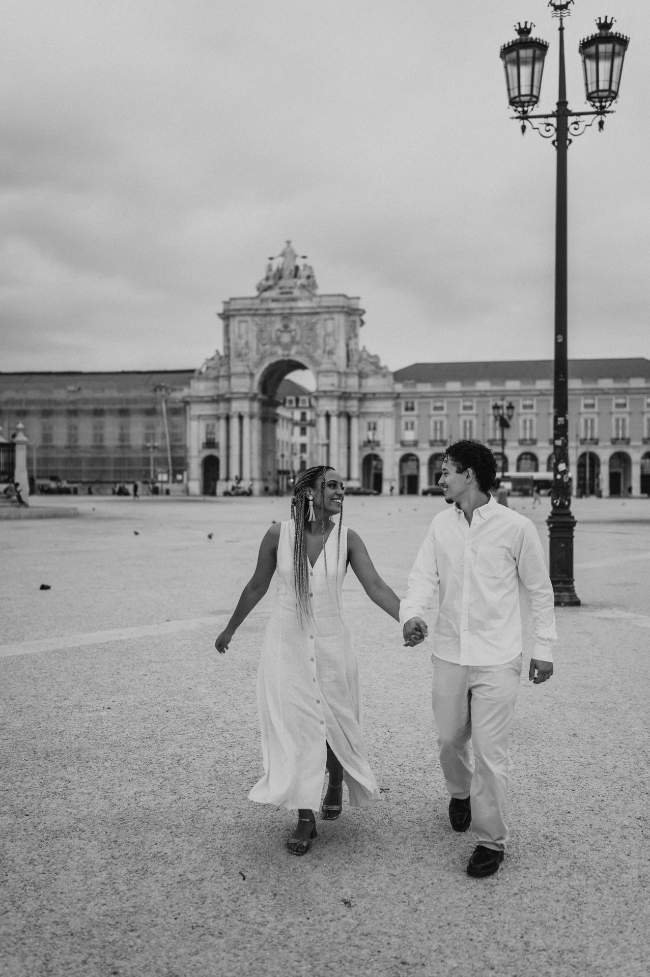 Black-and-white photograph: the couple walking hand in hand across Praça do Comércio with the monumental arch behind them during their couple photoshoot in Lisbon.