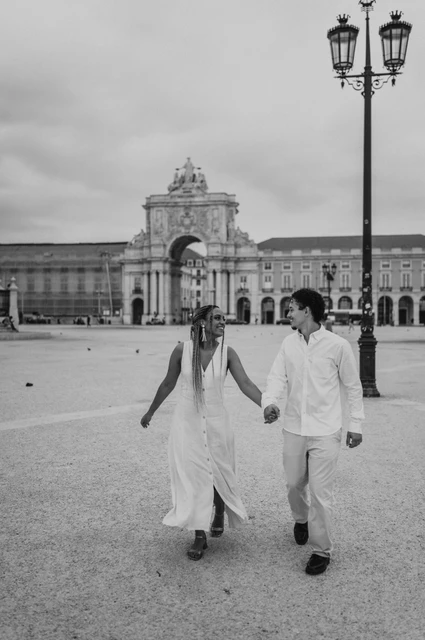 A walk across Praça do Comércio in black and white | Lisbon couple photoshoot Black-and-white photograph: the couple walking hand in hand across Praça do Comércio with the monumental arch behind them during their couple photoshoot in Lisbon.