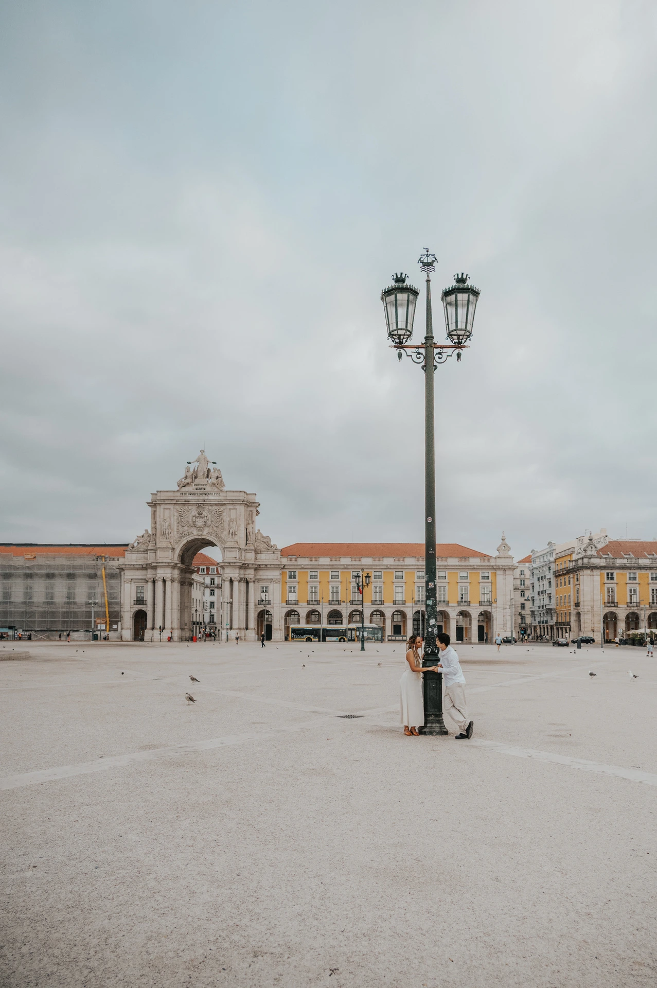 A wide view of the couple beside a tall street lamp in the open square at Praça do Comércio during their couple photoshoot in Lisbon.