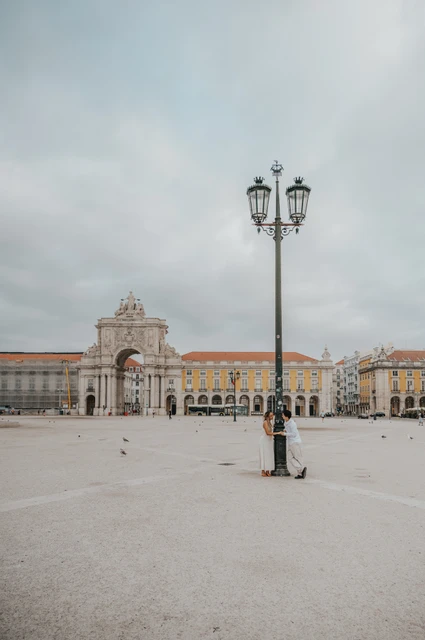 A wide view of the couple beside a tall street lamp in the open square at Praça do Comércio during their couple photoshoot in Lisbon.