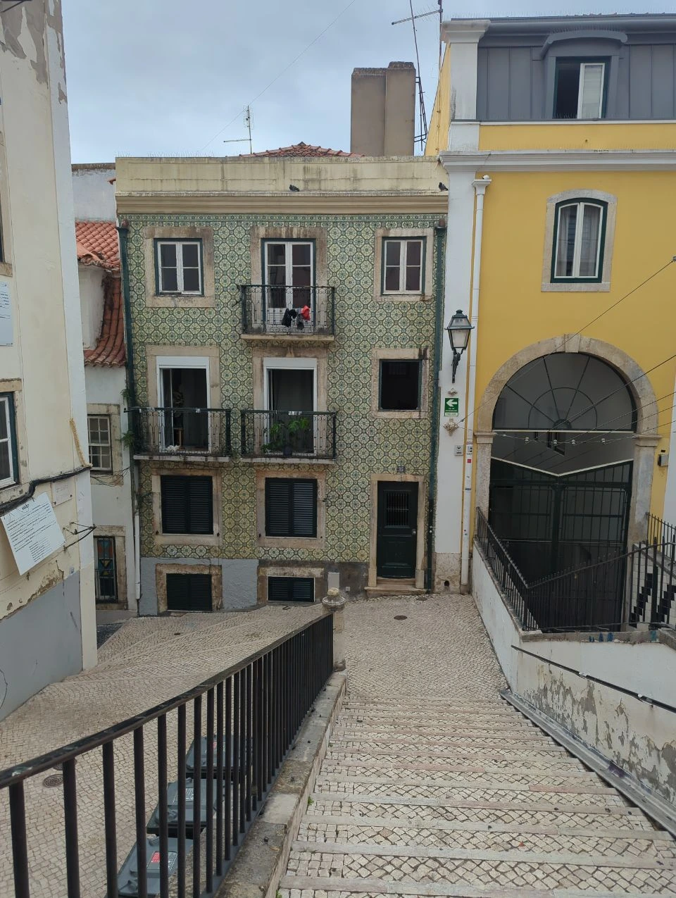 Lisbon Photoshoot Route - Steep Tiled Stair A steep patterned-stone stair descends between a green tiled Lisbon house, a yellow facade and black railings.