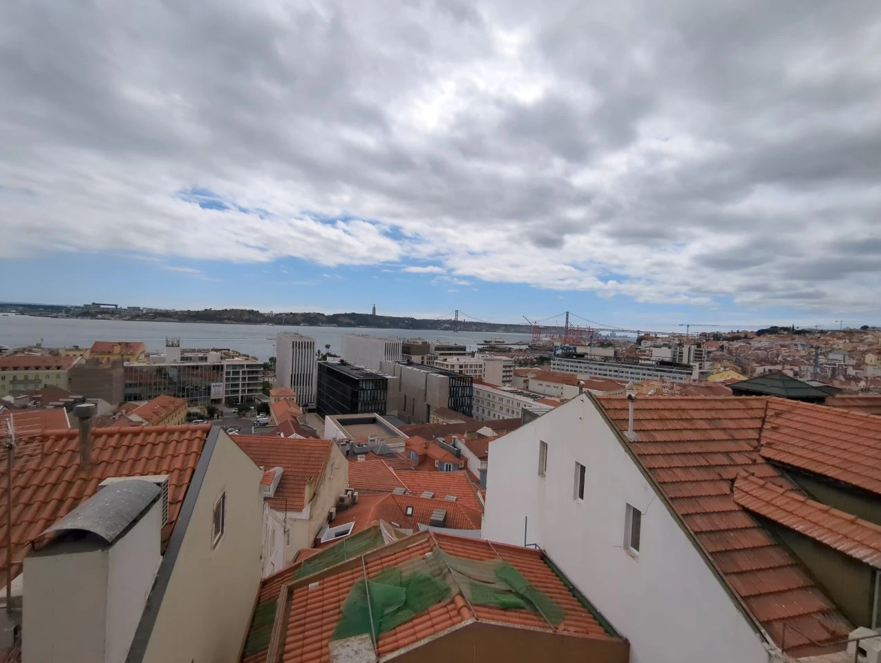 Red-roofed Lisbon stretches towards the Tagus and the suspension bridge beneath a broad, cloud-filled sky from an elevated viewpoint.