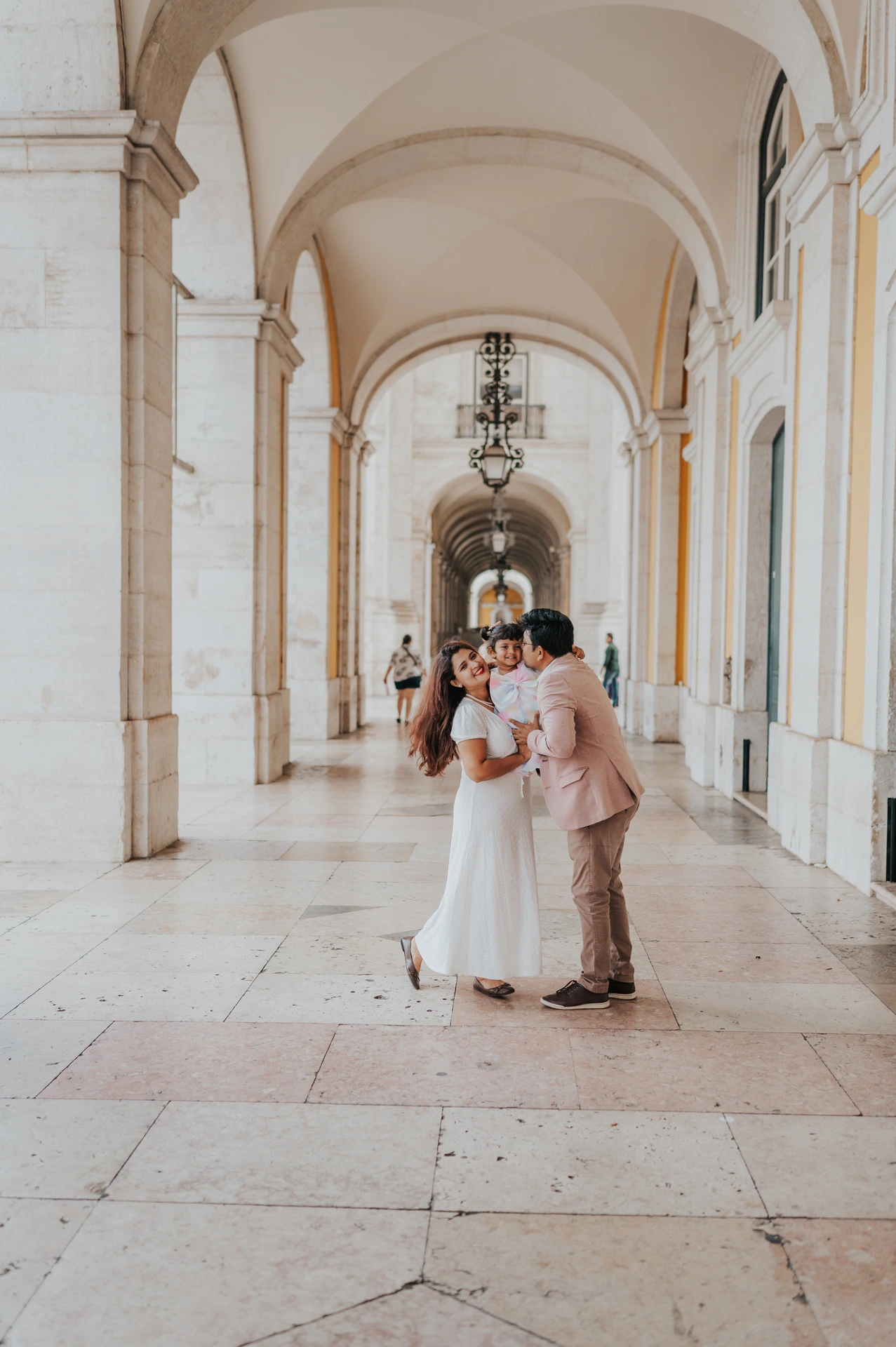 The parents holding their daughter close beneath the stone arcade at Praça do Comércio during their family birthday photoshoot in Lisbon.