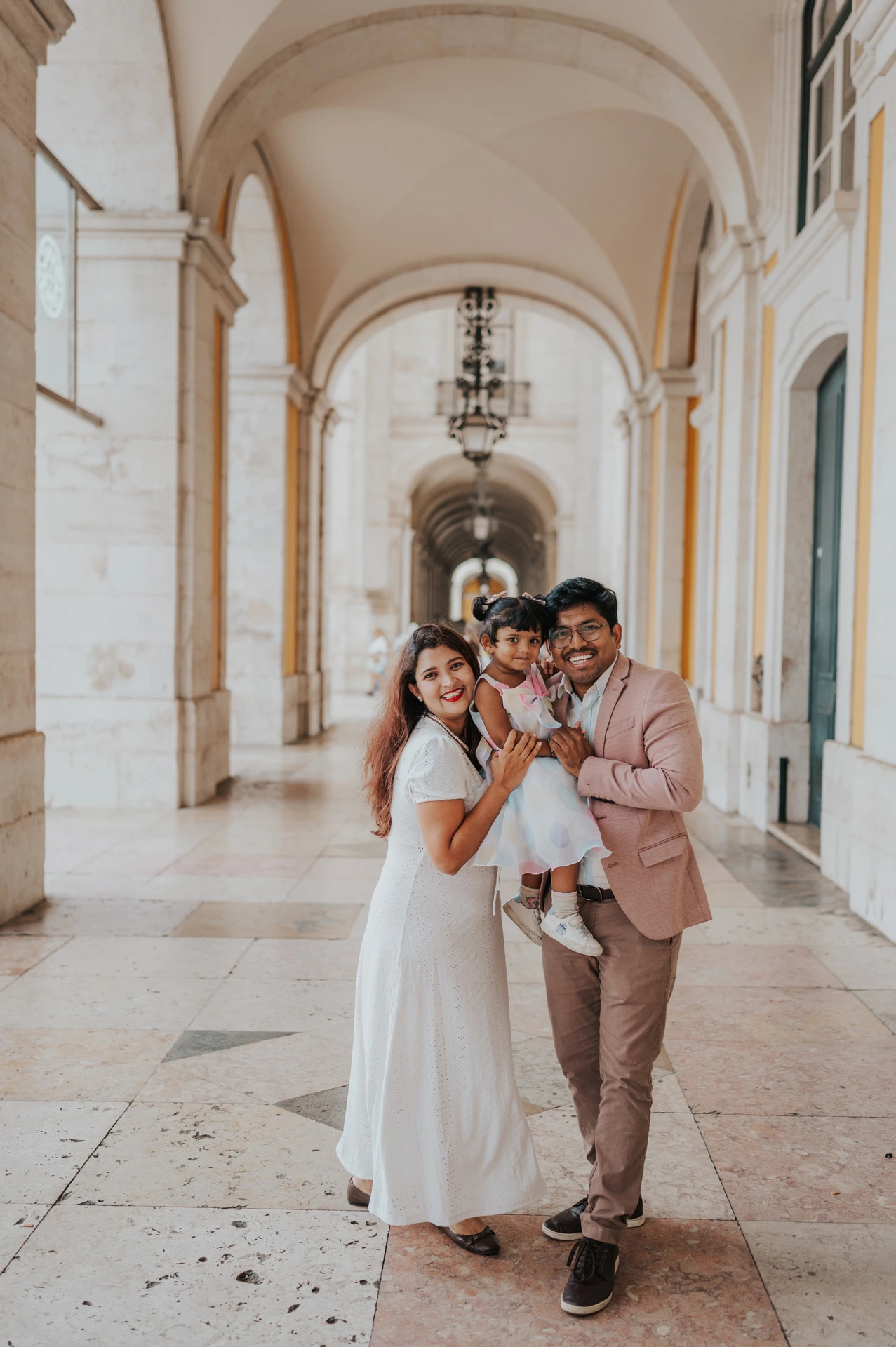 The parents holding their daughter close beneath the stone arcade at Praça do Comércio during their family birthday photoshoot in Lisbon.