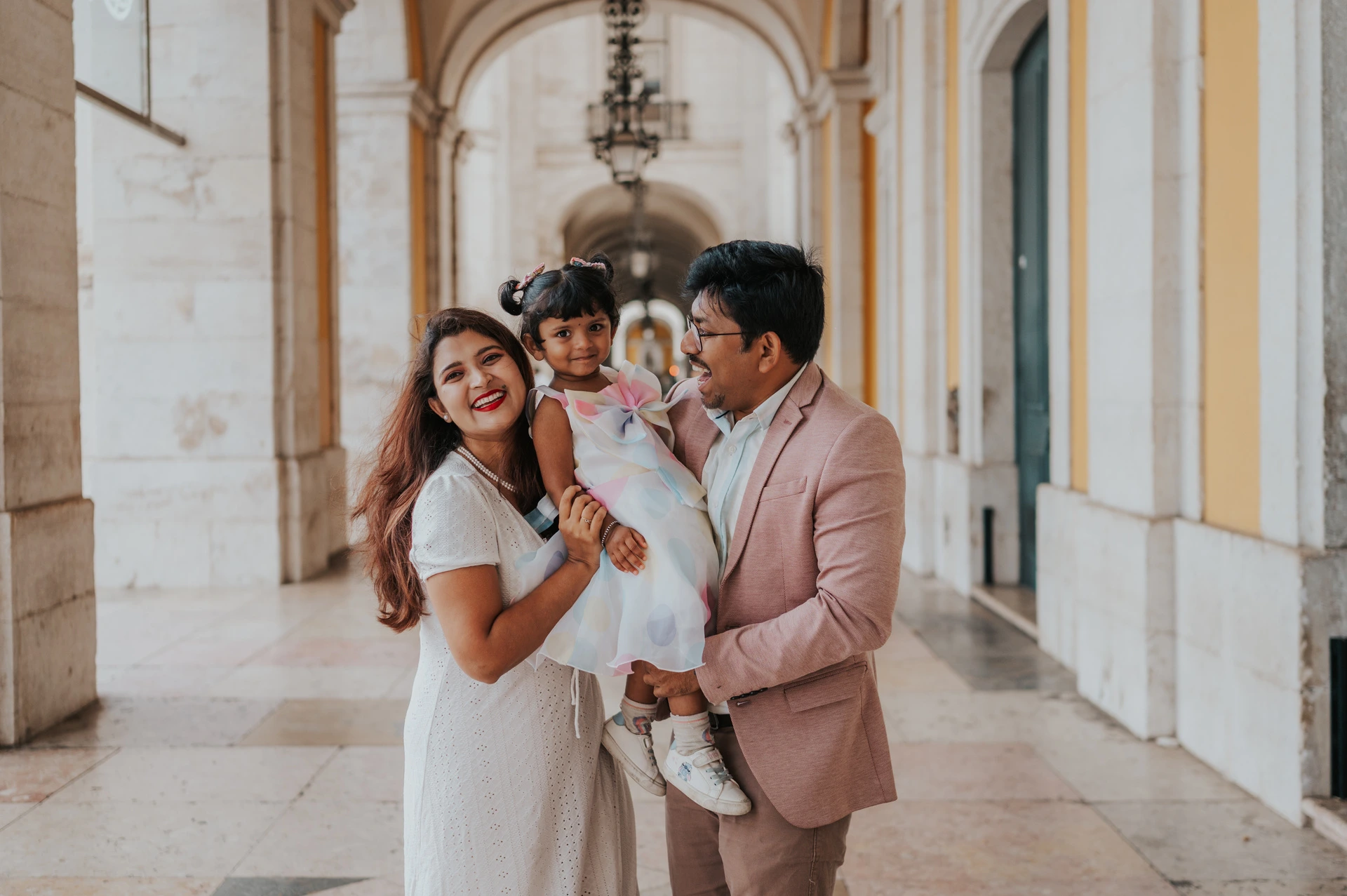 The parents holding their daughter close beneath the stone arcade at Praça do Comércio during their family birthday photoshoot in Lisbon.