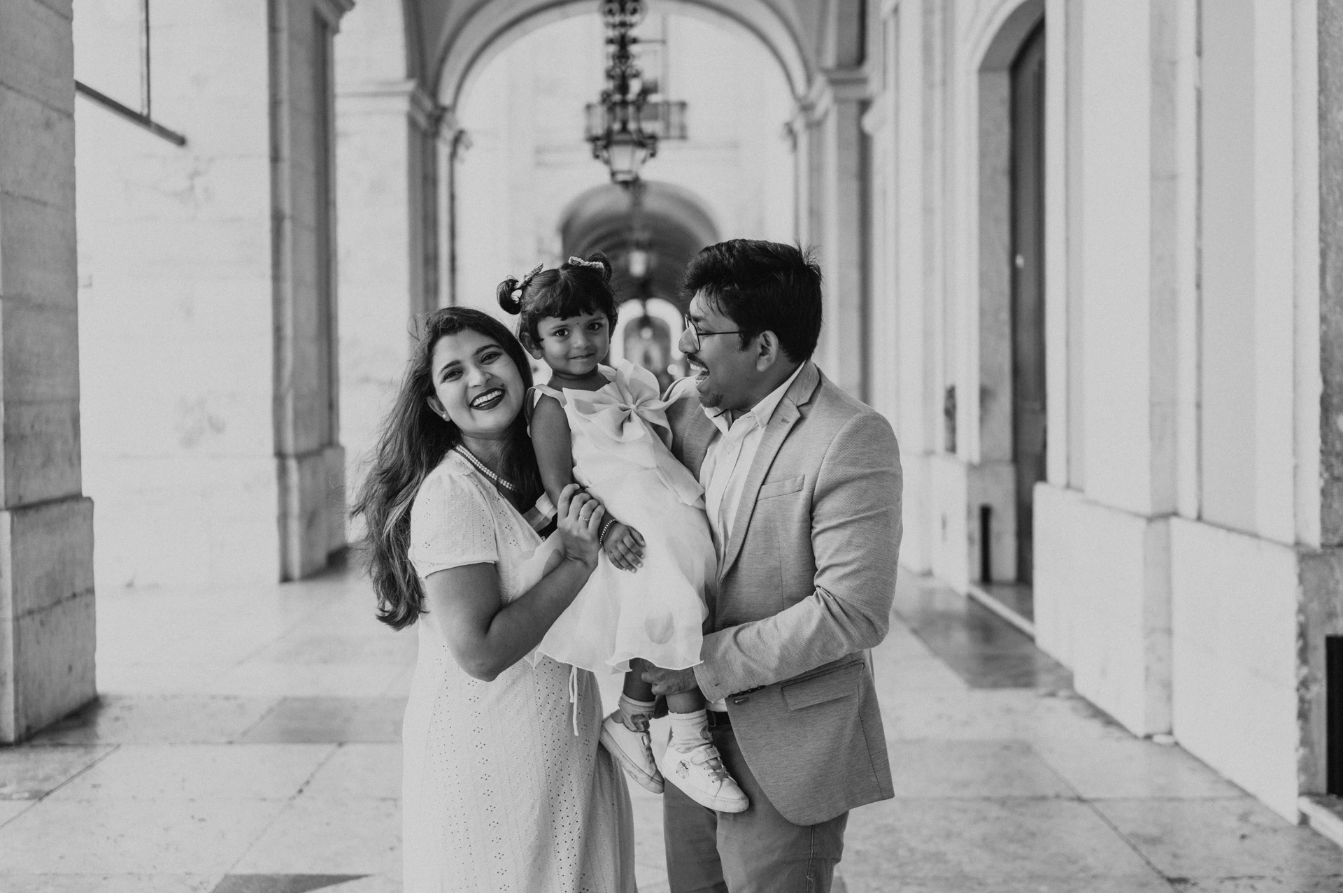 Black-and-white photograph: the parents holding their daughter close beneath the stone arcade at Praça do Comércio during their family birthday photoshoot in Lisbon.