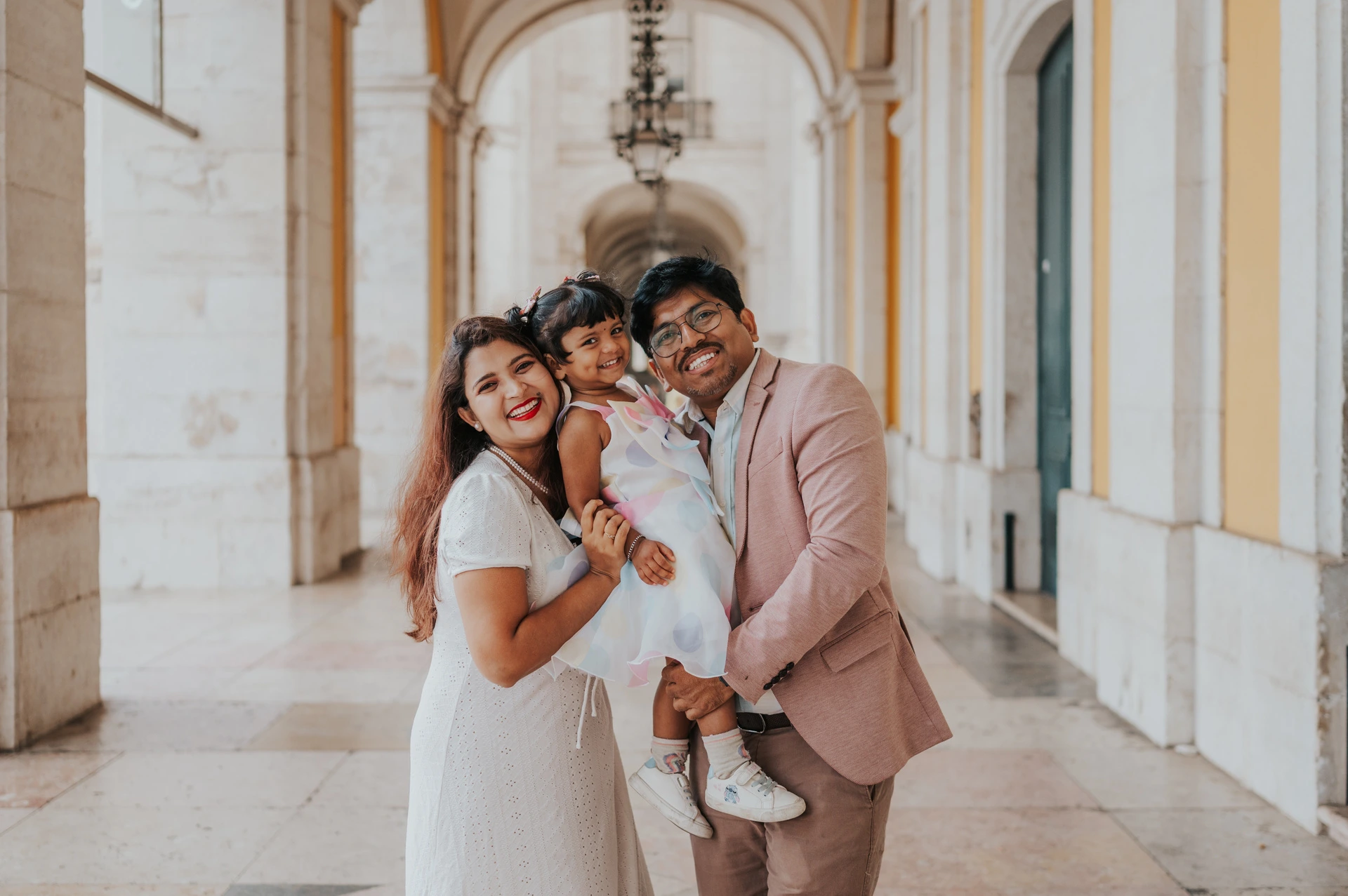 The parents holding their daughter close beneath the stone arcade at Praça do Comércio during their family birthday photoshoot in Lisbon.