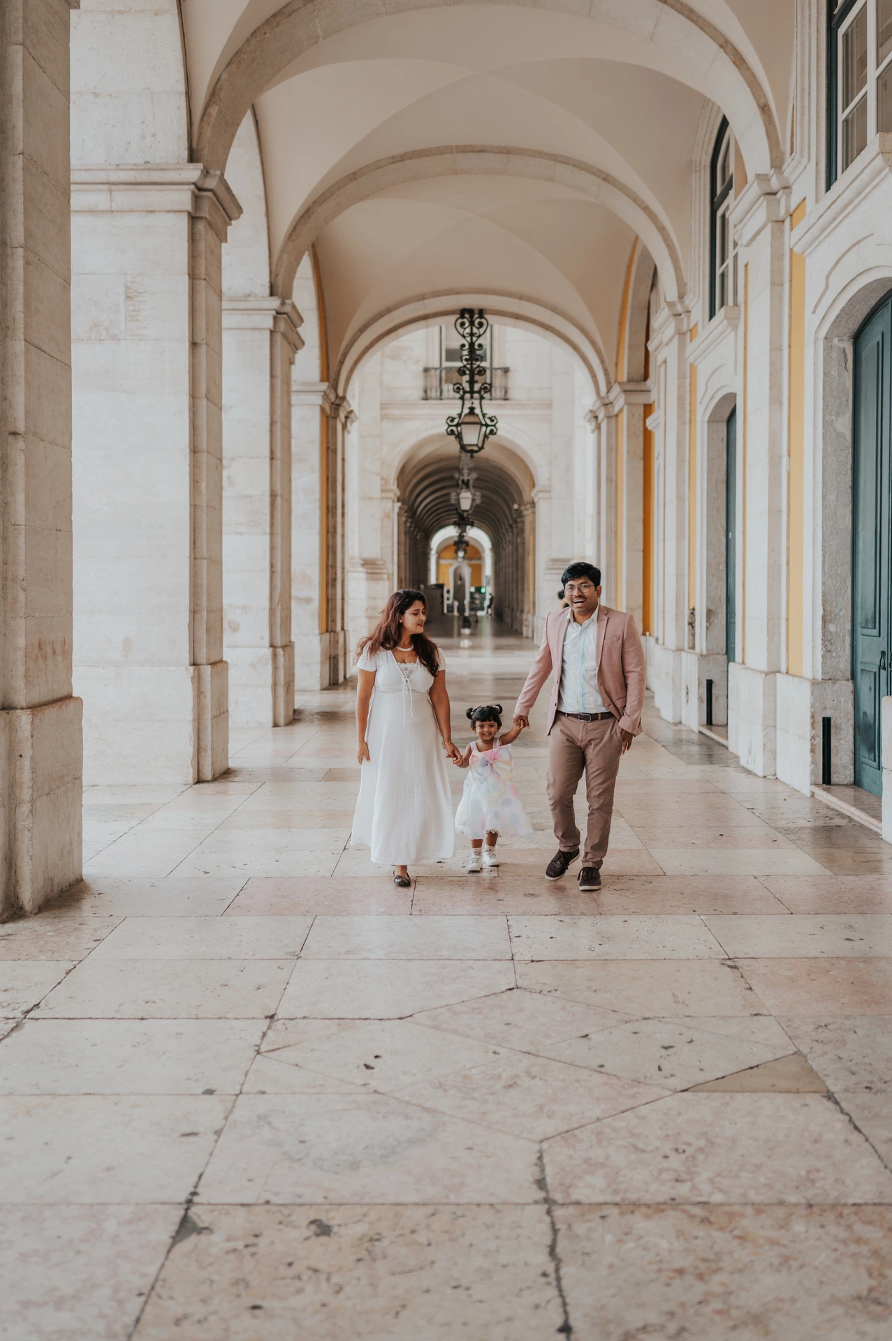 The parents holding their daughter’s hands as they walk together beneath the arcade during their family birthday photoshoot in Lisbon.