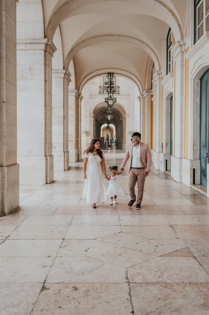 A playful family walk beneath the arches | Lisbon family birthday photoshoot The parents holding their daughter’s hands as they walk together beneath the arcade during their family birthday photoshoot in Lisbon.