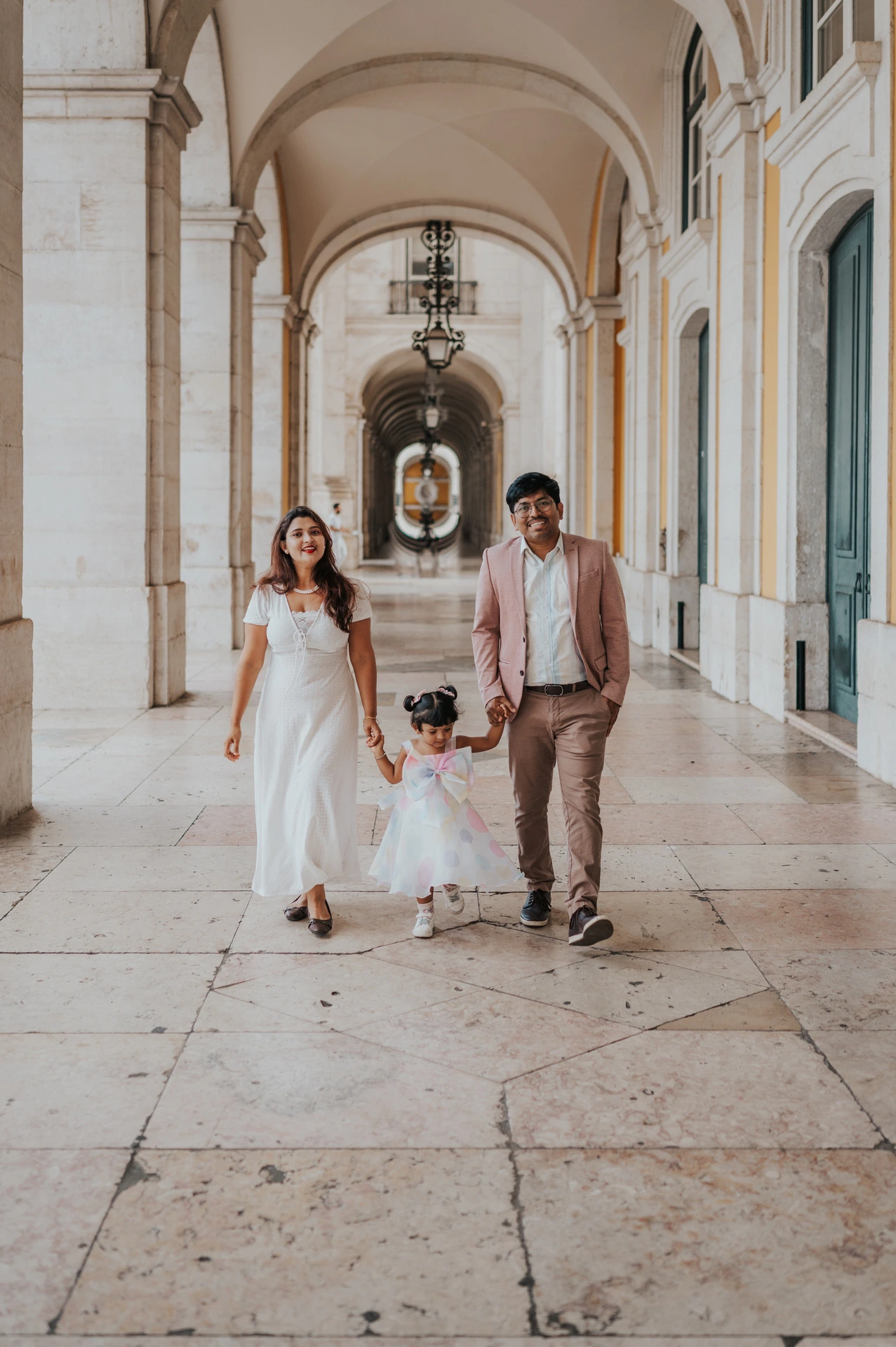 The parents holding their daughter’s hands as they walk together beneath the arcade during their family birthday photoshoot in Lisbon.