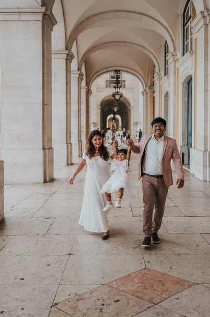 A playful family walk beneath the arches | Lisbon family birthday photoshoot The parents holding their daughter’s hands as they walk together beneath the arcade during their family birthday photoshoot in Lisbon.