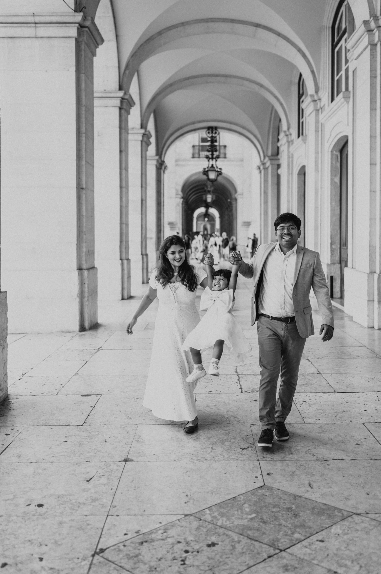 Black-and-white photograph: the parents holding their daughter’s hands as they walk together beneath the arcade during their family birthday photoshoot in Lisbon.
