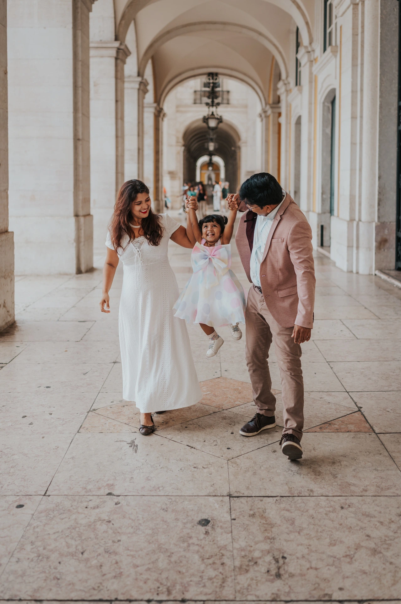 The parents holding their daughter’s hands as they walk together beneath the arcade during their family birthday photoshoot in Lisbon.
